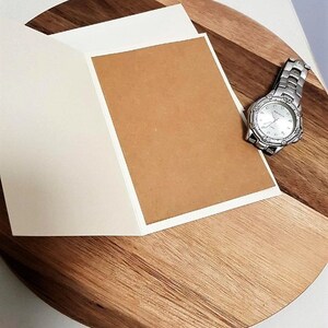 May include: An open greeting card with a blank brown interior and white border, placed on a wooden surface. A silver wristwatch with a white face is positioned nearby. The wooden surface has a natural wood grain pattern.