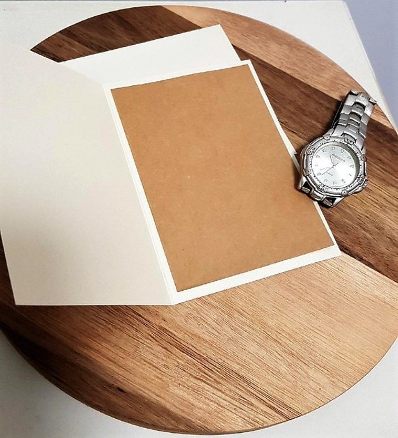 May include: An open greeting card with a blank brown interior and white border, placed on a wooden surface. A silver wristwatch with a white face is positioned nearby. The wooden surface has a natural wood grain pattern.