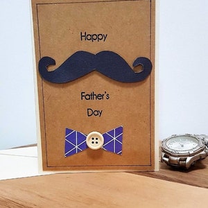 May include: A Father's Day greeting card with a brown background, featuring a black mustache and a blue bow tie with a white button. The words "Happy Father's Day" are printed on the card.