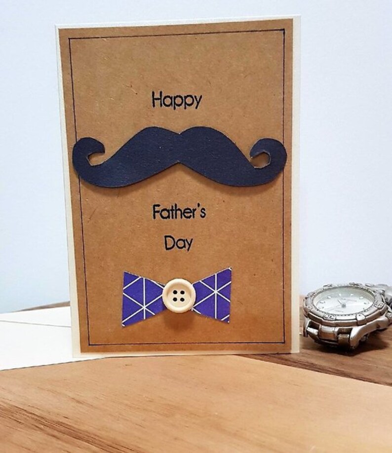 May include: A Father's Day greeting card with a brown background, featuring a black mustache and a blue bow tie with a white button. The words "Happy Father's Day" are printed on the card.