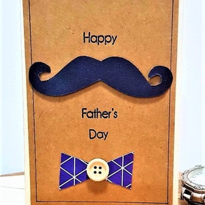 May include: A handmade Father's Day card with a brown background. The card features a dark blue mustache, the words "Happy Father's Day", and a blue bow tie with a white geometric pattern. A button is in the center of the bow tie.