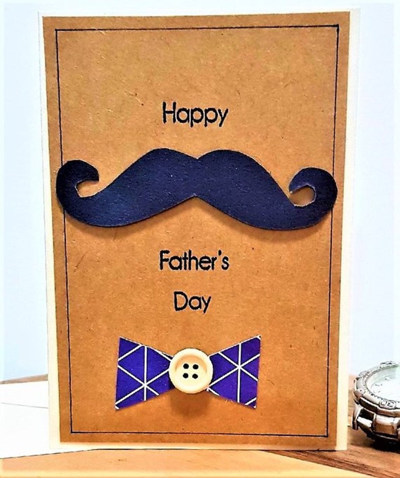 May include: A handmade Father's Day card with a brown background. The card features a dark blue mustache, the words "Happy Father's Day", and a blue bow tie with a white geometric pattern. A button is in the center of the bow tie.