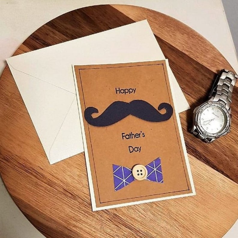 May include: A Father's Day card with a brown background, a black mustache, and a blue bow tie with a button. The card reads "Happy Father's Day." An envelope and a silver wristwatch are also present.