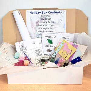 May include: A white box filled with craft supplies for children. The box includes paint, play dough, colouring pages, recipes to cook, lacing cards, and collage materials. The box is labelled "Holiday Box Contents".