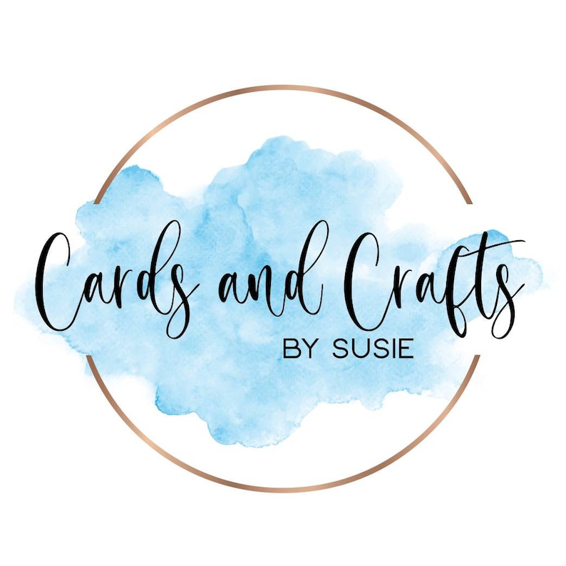 May include: Logo for "Cards and Crafts by Susie". The text is in a black, elegant script, set against a light blue watercolor cloud. A rose gold circle frames the design, creating a sophisticated look.