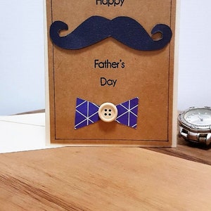 May include: A Father's Day greeting card with a navy blue mustache and bow tie. The bow tie is purple with white geometric patterns and a button center. The words "Happy Father's Day" are printed on the card.