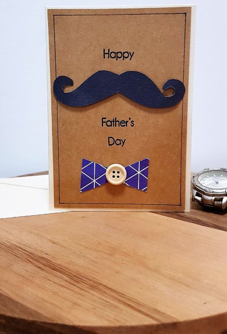 May include: A Father's Day greeting card with a navy blue mustache and bow tie. The bow tie is purple with white geometric patterns and a button center. The words "Happy Father's Day" are printed on the card.