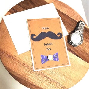 May include: A Father's Day card with a brown background, featuring a black mustache and a purple bow tie. The words "Happy Father's Day" are printed on the card. A silver wristwatch is also visible.
