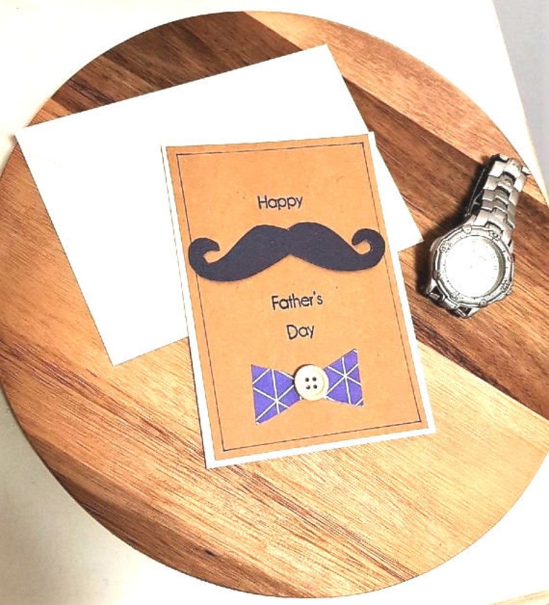 May include: A Father's Day card with a brown background, featuring a black mustache and a purple bow tie. The words "Happy Father's Day" are printed on the card. A silver wristwatch is also visible.