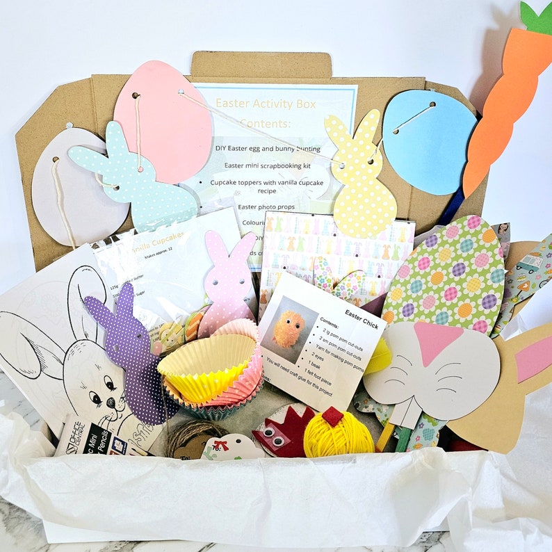 May include: A white box filled with craft supplies for an Easter activity box. The box includes paper cutouts of bunnies, eggs, and carrots, cupcake liners, yarn, and a chick craft kit. The box also includes instructions for making Easter crafts.