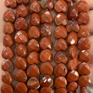 May include: Close-up of a strand of heart-shaped, faceted red jasper beads. The beads have a warm, earthy tone with variations in color and pattern. The facets catch the light, creating a sparkling effect. Ideal for jewelry making.