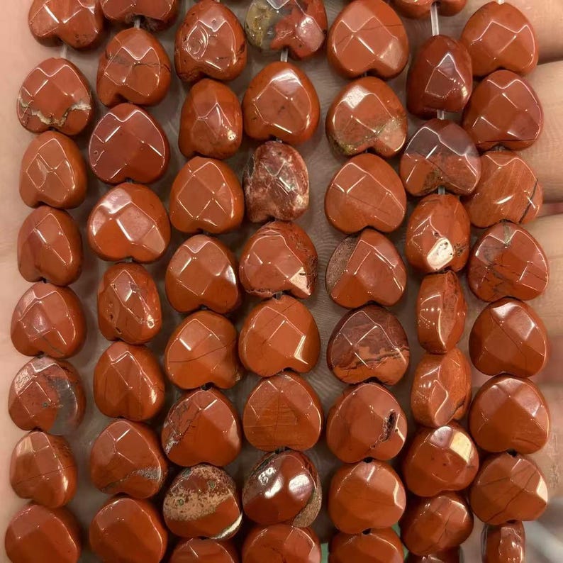 May include: Close-up of a strand of heart-shaped, faceted red jasper beads. The beads have a warm, earthy tone with variations in color and pattern. The facets catch the light, creating a sparkling effect. Ideal for jewelry making.
