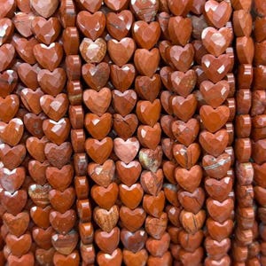 May include: Strands of heart-shaped beads in shades of red-orange. The beads have a faceted surface, catching the light. The beads are strung together, creating a textured and visually appealing pattern. The beads are made of a natural stone.