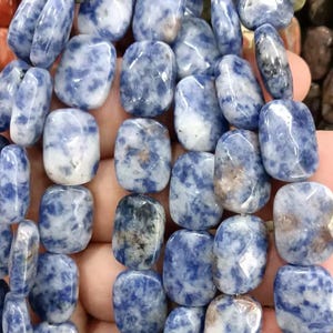 May include: Close-up of a strand of sodalite beads. The beads are a mix of blue and white, with a polished, smooth surface. The beads are oval-shaped and strung together, creating a necklace or bracelet.
