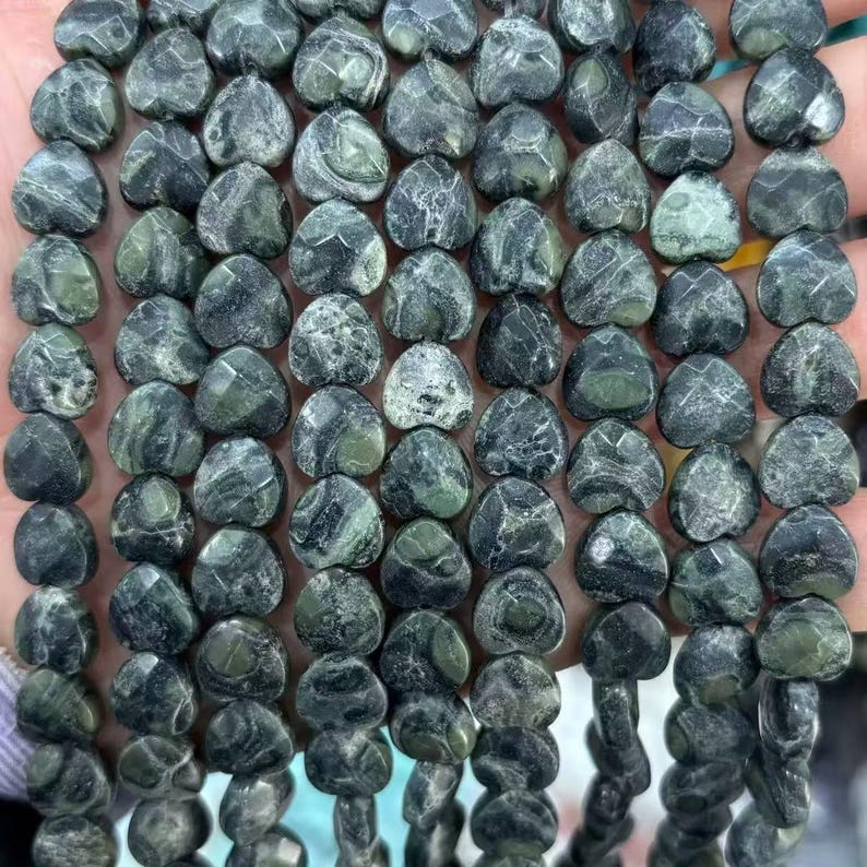 May include: Strands of heart-shaped beads in shades of green and gray. The beads have a marbled appearance, with variations in color and texture. The beads are strung together, creating a necklace or bracelet.