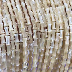 Puede incluir: Una hebra de cuentas de concha de color blanco y crema con un acabado ligeramente iridiscente. Las cuentas tienen forma de pequeños cilindros alargados con una ligera forma de reloj de arena.