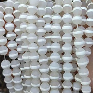Può includere: Primo piano di fili di perline bianche smerigliate, ciascuna con una singola striscia bianca. Le perline sono sferiche e infilate insieme, formando un motivo strutturato. Queste perline di colore neutro possono essere utilizzate per la creazione di gioielli.