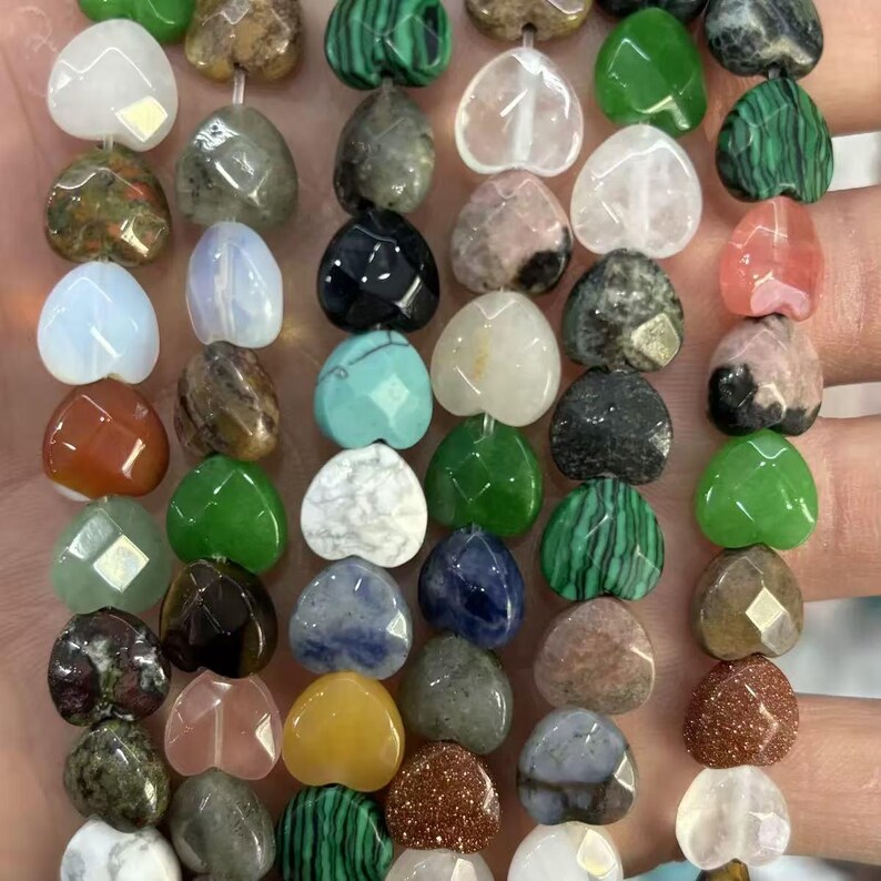May include: A collection of heart-shaped gemstone beads in various colors and textures. The beads are strung together, showcasing a variety of materials including green malachite, clear quartz, and speckled stones. The beads are approximately 0.5 inches in size.