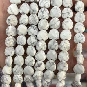 May include: A strand of white heart-shaped beads with gray marbling. The beads are smooth and polished, strung together, and ready for jewelry making. The hearts are approximately 1 cm in size.