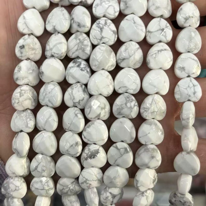 May include: A strand of white heart-shaped beads with gray marbling. The beads are smooth and polished, strung together, and ready for jewelry making. The hearts are approximately 1 cm in size.