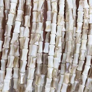 Peut inclure: Un brin de perles de coquillage blanches et beiges de forme cylindrique. Les perles ont une finition lisse et polie et un léger éclat iridescent.