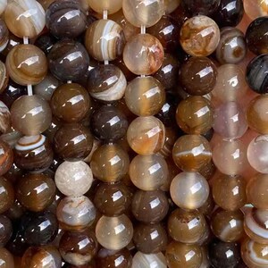 Puede incluir: Primer plano de un collar de cuentas de ágata pulidas. Las cuentas son una mezcla de colores marrón, beige y crema, con patrones de bandas naturales. Las cuentas redondas están ensartadas, creando una superficie texturizada. Las cuentas tienen aproximadamente 1 cm de diámetro.