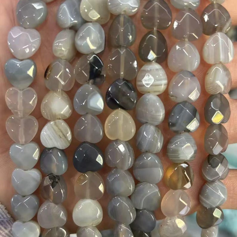 May include: Strands of heart-shaped beads in various shades of gray, beige, and brown. The beads have a faceted or smooth surface, with some showing natural stone patterns. The beads are strung together, ready for jewelry making.