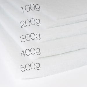 Könnte beinhalten: Nahaufnahme von mehreren weißen, gefalteten Stoffquadraten, die jeweils mit einem Gewicht in Gramm beschriftet sind: 100g, 200g, 300g, 400g und 500g. Der Stoff wirkt weich und flauschig.