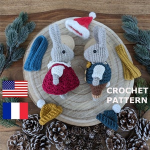 May include: Crochet pattern for two grey bunny amigurumi toys wearing Santa hats. The bunny on the left is wearing a red dress and the bunny on the right is wearing a blue waistcoat. The pattern is displayed on a wooden surface with pine cones and other decorations.