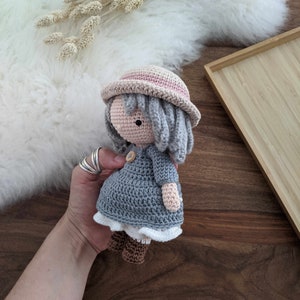 May include: A crocheted doll wearing a gray dress, a pink hat, and brown boots. The doll has gray yarn hair and a button nose.