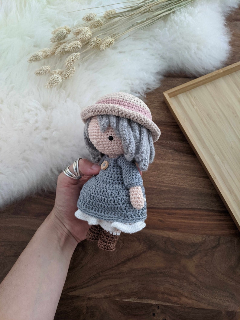 May include: A crocheted doll wearing a gray dress, a pink hat, and brown boots. The doll has gray yarn hair and a button nose.
