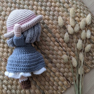 May include: A crocheted doll wearing a pink and white hat and a blue dress. The doll is standing on a woven mat with dried grass.