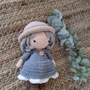 May include: A crocheted doll wearing a gray dress, a pink and white hat, and brown boots. The doll is sitting on a woven rug with a green leafy plant in the background.