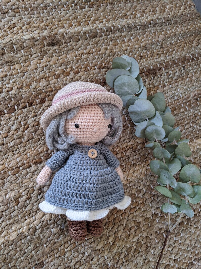 May include: A crocheted doll wearing a gray dress, a pink and white hat, and brown boots. The doll is sitting on a woven rug with a green leafy plant in the background.