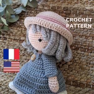 Op de afbeelding: Haakpatroon voor een pop met een roze en beige hoed, grijs haar en een blauwe jurk. De pop zit op een geweven oppervlak. De tekst "CROCHET PATTERN" is zichtbaar in de afbeelding.