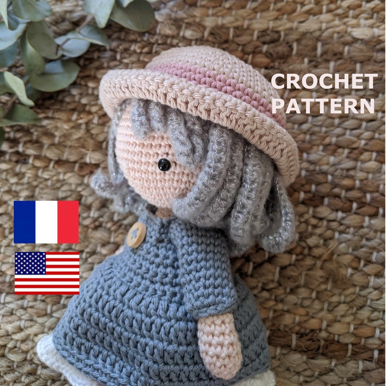 May include: Crochet pattern for a doll with a pink and beige hat, gray hair, and a blue dress. The doll is sitting on a woven surface. The text "CROCHET PATTERN" is visible in the image.