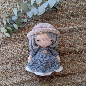 May include: A crocheted doll wearing a blue dress, a pink and white hat, and brown shoes. The doll is sitting on a woven rug.
