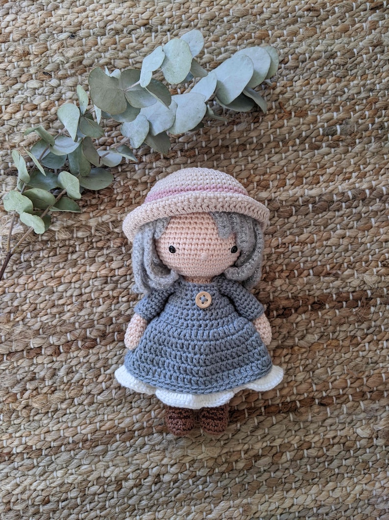 May include: A crocheted doll wearing a blue dress, a pink and white hat, and brown shoes. The doll is sitting on a woven rug.