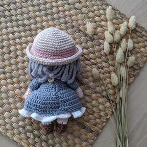 May include: A crocheted doll wearing a pink and beige hat, a gray coat, and white shoes. The doll is sitting on a woven mat.