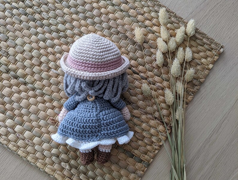 May include: A crocheted doll wearing a pink and beige hat, a gray coat, and white shoes. The doll is sitting on a woven mat.