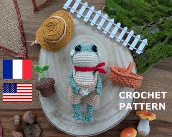 May include: A crocheted frog doll in dungarees and a red scarf, with a small hat, basket, and plant. The frog is light blue and beige. The image also includes a white picket fence and the words "CROCHET PATTERN".