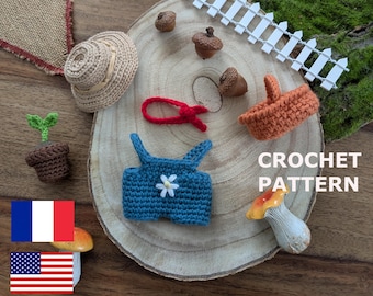 May include: A selection of miniature crochet items, including a straw hat, blue overalls with a flower, a basket, a plant, and acorns. The text "CROCHET PATTERN" is visible. The items are arranged on a wooden slice.