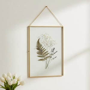 Puede incluir: Un marco de pared rectangular con un borde fino de metal dorado y una delicada cadena dorada para colgar. Dentro del cristal, una hoja de helecho verde prensada y pequeñas flores secas blancas están dispuestas sobre un fondo blanco.