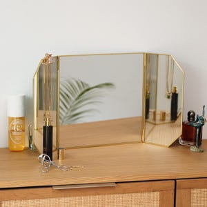 May include: A gold-toned, three-sided vanity mirror with bevelled edges. The mirror is sitting on a wooden surface with a variety of items, including a bottle of perfume, a bottle of lotion, and a pair of eyelash curlers.