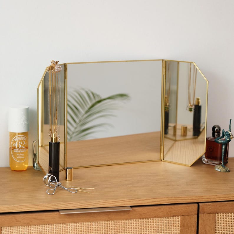 May include: A gold-toned, three-sided vanity mirror with bevelled edges. The mirror is sitting on a wooden surface with a variety of items, including a bottle of perfume, a bottle of lotion, and a pair of eyelash curlers.