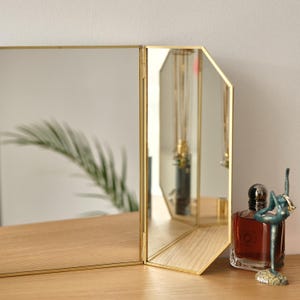 May include: A three-sided mirror with a gold frame and bevelled edges. The mirror is standing on a wooden surface with a bottle of perfume and a decorative figurine in front of it.
