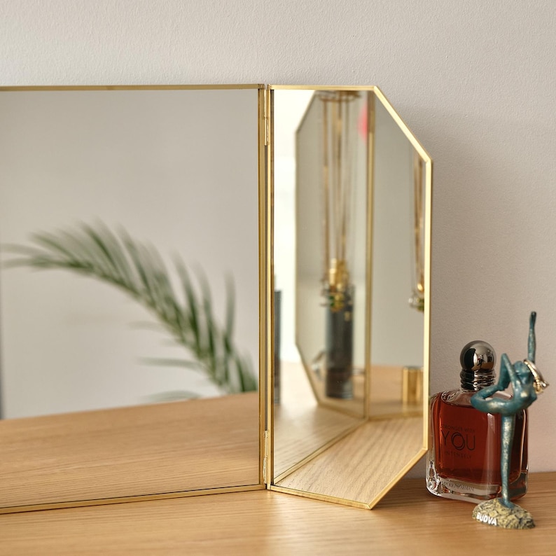 May include: A three-sided mirror with a gold frame and bevelled edges. The mirror is standing on a wooden surface with a bottle of perfume and a decorative figurine in front of it.