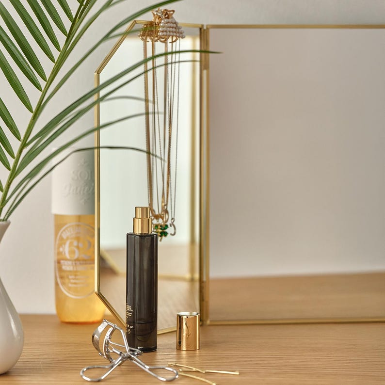 May include: A gold-framed vanity mirror with a gold-coloured bottle of perfume and a gold-coloured eyelash curler on a wooden surface. The mirror has a decorative design and is reflecting a necklace and other jewellery.