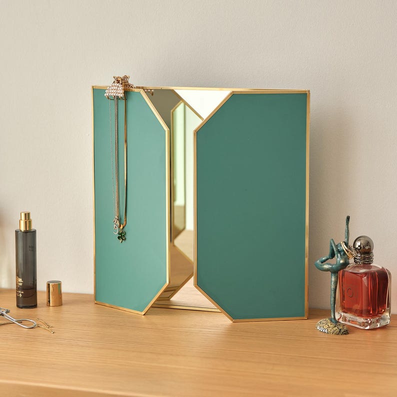 May include: A teal green and gold framed tri-fold mirror with a bevelled edge. The mirror is on a wooden surface with a perfume bottle, a hair clip, and a small decorative figurine.