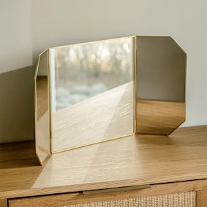 May include: A gold-toned, three-sided vanity mirror with a bevelled edge. The mirror is sitting on a wooden surface.
